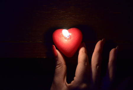 Women's hand holding a burning red candle in the dark lightの写真素材