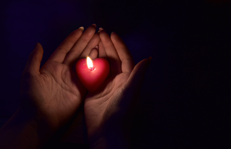 Women's hand holding a burning red candle in the dark lightの写真素材