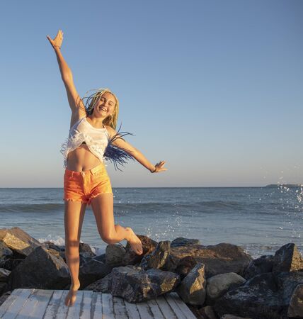 Senegalese girl with pigtails jumps on the wooden jetty on the sea. The waves are hitting the rocks and the spray is flying in different directions. Vacation.の写真素材