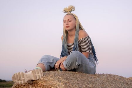 Girl with Senegalese pigtails sitting on a haystack and looking into the distance. Braids twisted on her head in a bun. Illuminated by the setting sun.の写真素材