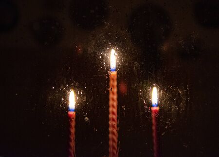 Three blurred red candles burning in the darkness outside the window, where raindrops are dripping.の写真素材