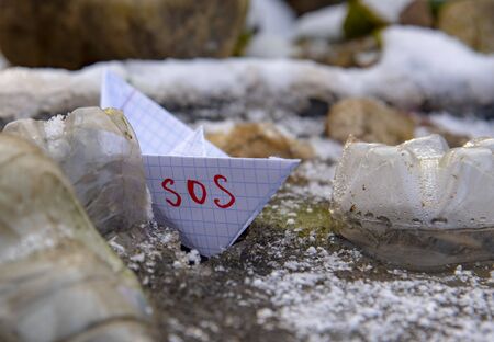 The paper boat marked SOS was stuck among the plastic bottles frozen in the water. Conceptual photography.の写真素材