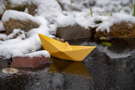 Yellow bright paper boat stuck in the ice, on a frozen puddle.
