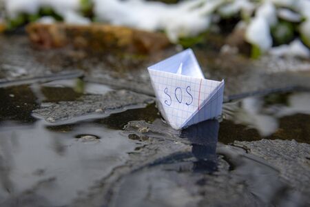 A paper boat made of a checkered notebook with the inscription SOS stuck in the ice.の写真素材
