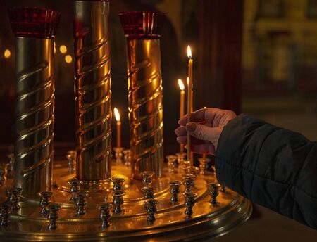 The hand of an elderly woman puts a lighted candle to the icon. On dark background.の写真素材