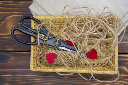 A wicker basket with scissors, coarse thread, and red felt hearts stands on Kraft paper against a dark wooden background.の写真素材