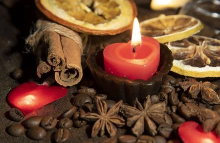 A burning candle in the shape of a heart, various spices, dried oranges and small bright meringues on a wooden table top.の写真素材