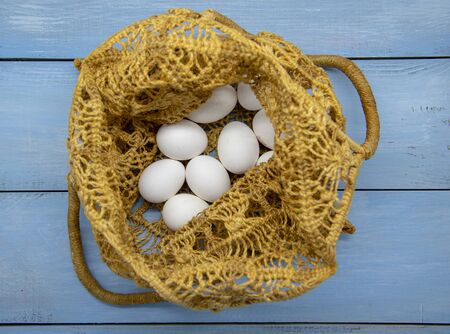 White eggs in a wicker bag made of eco-friendly materials on a blue wooden tabletop.の写真素材
