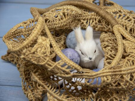 Toy bunny and Easter painted eggs in a wicker bag from environmentally friendly materials on a blue...