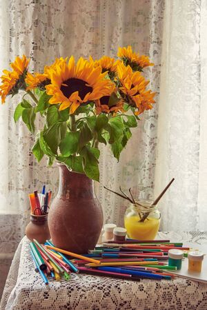 Bright sunflowers in a clay jug on the table by the window. Pencils, paints, and brushes are scattered around. Creative mess.の写真素材