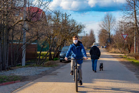 Russia, Tuchkovo village, April 2020. A young man in a protective medical mask and rubber gloves rides a Bicycle along a village street. The epidemic of the coronavirus, stay home.のeditorial素材