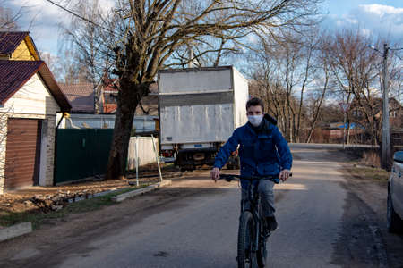 Russia, Tuchkovo village, April 2020. A young man in a protective medical mask and rubber gloves rides a Bicycle along a village street. The epidemic of the coronavirus, stay home.のeditorial素材