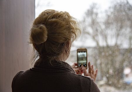 An elderly woman photographs an unexpected spring snowfall on her phone through a doorway during a coronavirus epidemic.の写真素材