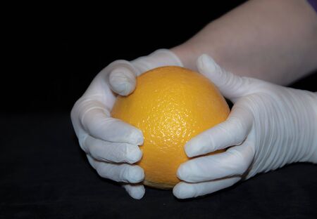 The hands of an elderly woman in white rubber gloves hold a large bright orange orange. On black background.の写真素材