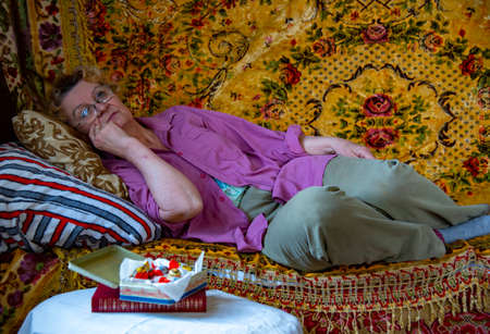 A tired old woman in a mans shirt is lying on a sofa covered with vintage carpets.のeditorial素材