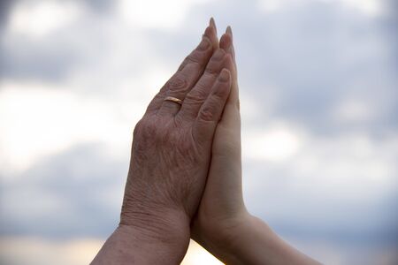 The hand of an elderly woman and the hand of a teenage girl on the background of a cloudy sky.