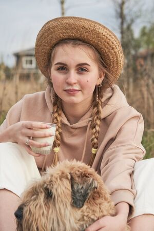 A teenage girl with pigtails and a hat with a glass of milk in her hands and a milky mustache hugs a dog.の写真素材