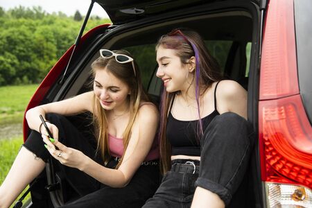 Two female friends with long hair, a blonde and a brunette, sit in the trunk of a car and look at...