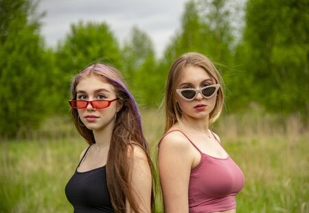 Two teenage girls with long loose hair and sunglasses pose as models against the backdrop of nature.