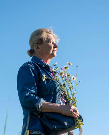 An elderly woman of 70 years, illuminated by sunlight, poses against the blue sky with a bouquet of daisies.の写真素材