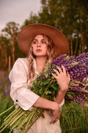 A girl in a white shirt and a straw hat with a bouquet of flowers poses in a Lupin field.の写真素材