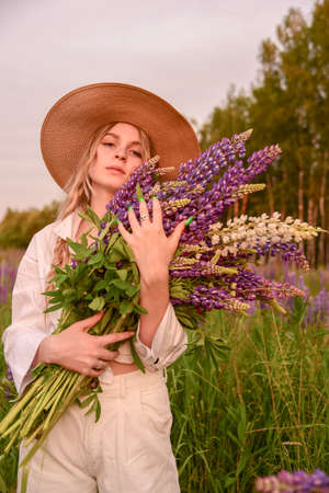 A girl in a white shirt and a straw hat with a bouquet of flowers poses in a Lupin field.の写真素材