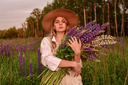 A girl in a white shirt and a straw hat with a bouquet of flowers poses in a Lupin field.の写真素材
