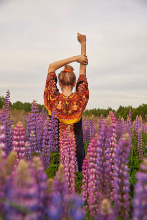 A girl in a dress with ethnic ornaments stands in a lupine field with her hands up and her back to the camera.の写真素材