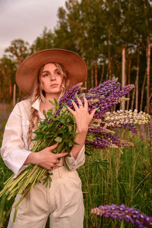 A girl in a white shirt and a straw hat with a bouquet of flowers poses in a Lupin field.の写真素材
