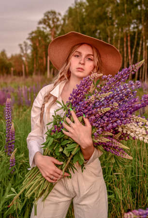 A girl in a white shirt and a straw hat with a bouquet of flowers poses in a Lupin field.の写真素材