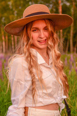 A flirtatious beautiful teenage girl with long blonde hair, wearing a straw hat and white clothes, poses against a background of blooming lupines. Photo session in nature.の写真素材
