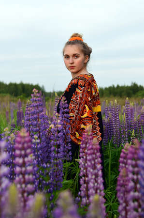 A beautiful young girl with blonde hair tied in a bun, in an ethnic dress, posing against a background of blooming lupines. Photo session in nature.の写真素材