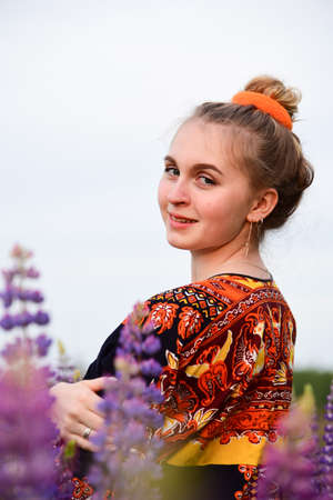 A beautiful young girl with blonde hair tied in a bun, in an ethnic dress, posing against a background of blooming lupines. Photo session in nature.の写真素材