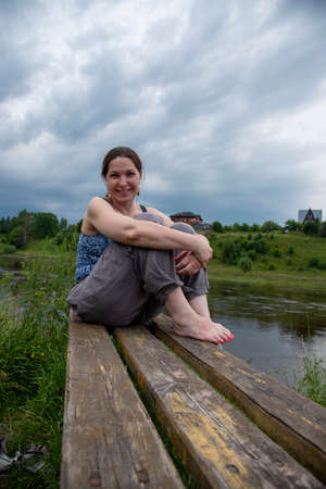 A middle-aged woman sits on a wooden bench on the river Bank on a cloudy summer day.の写真素材