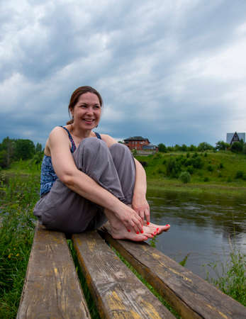 A middle-aged woman sits on a wooden bench on the river Bank on a cloudy summer day.の写真素材