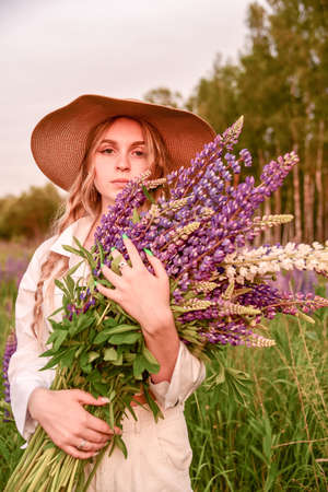 A young beautiful girl in a white shirt and a wide-brimmed straw hat with a bouquet of flowers poses in a Lupine field.の写真素材