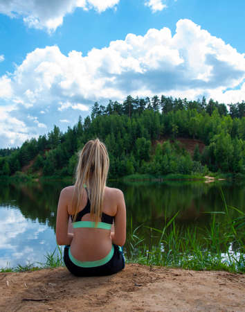 A girl with long blond hair in a sports top sits on the lake with her back to the camera.の写真素材