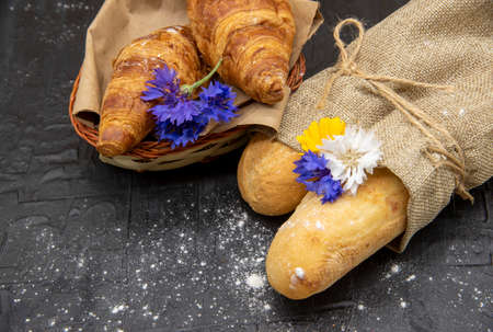 Baguettes wrapped in coarse linen and a basket of croissants decorated with flowers on a black slate background.の写真素材
