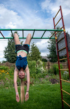 A girl with long blond hair and sports clothes is hanging upside down on a horizontal bar.の写真素材