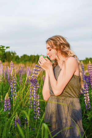 A blonde girl with long wavy hair in an elegant gray dress with an open shoulder poses in lupine fields.の写真素材