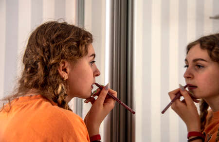 A teenage girl with pigtails stands in front of a mirror and paints her lips with a cosmetic pencil.の写真素材