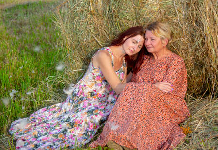 Two women, a mother and daughter, in bright dresses, sit side by side, leaning against a haystack.の写真素材