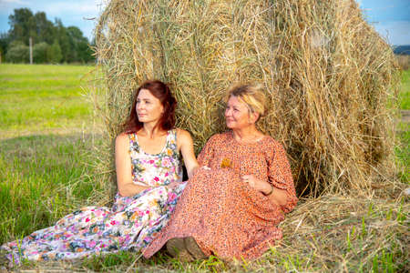 Two women, a mother and daughter, in bright dresses, sit side by side, leaning against a haystack.の写真素材