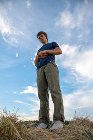 A young man stands on a haystack against a blue cloudy sky and looks at the camera.の写真素材
