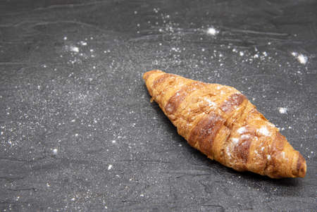One croissant, sprinkled with flour, lies on a black slate background.の写真素材