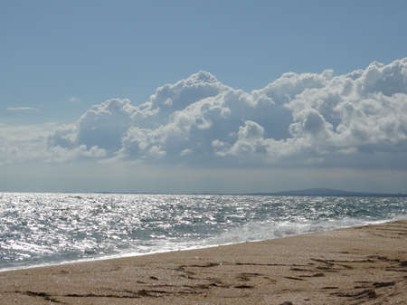 An empty sandy beach and a foaming sea under a cloudy sky. The sea is silver under the sun.の写真素材