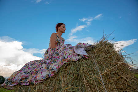 Photo of a red-haired middle-aged woman in a long summer sundress, sitting on a haystack.の写真素材