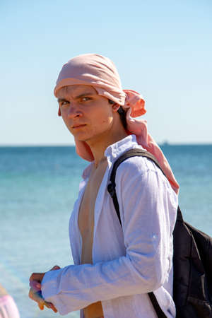 A gloomy young man in a bandana and a white shirt, with a backpack, against the sea.の写真素材