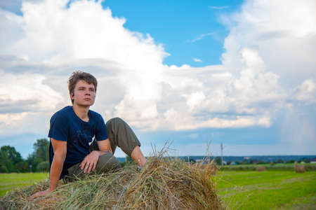 A handsome young man is sitting on a haystack in the fields against a blue cloudy sky.の写真素材