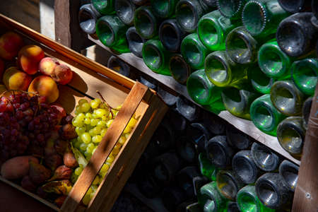 Dusty wine bottles are stacked on racks, and a wooden crate of juicy fruit stands next to them.の写真素材